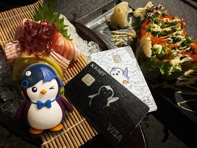 The image features a penguin figurine sitting next to two credit cards - one Visa and one Mastercard. The penguin figurine has a blue bowtie around its neck, adding a playful touch to the scene. The cards are placed on top of a sushi platter that is accompanied by chopsticks and other food items such as fish and vegetables. The post includes text about penguins and credit card holders, suggesting an interesting connection between these two elements in the image.