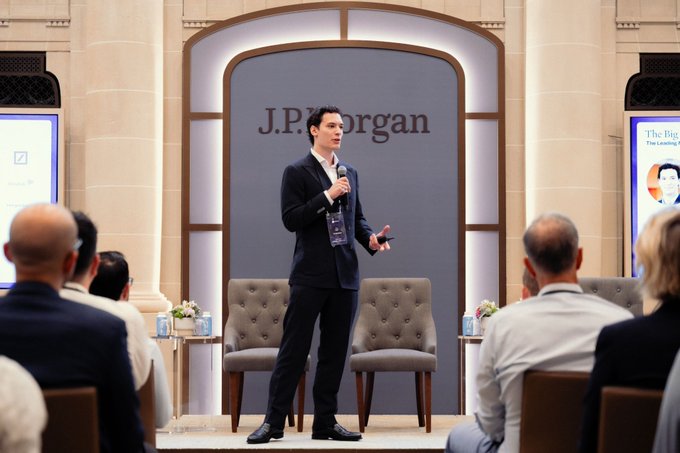 A speaker addresses attendees at a J.P. Morgan-hosted Corporate Breakfast in Paris, with the "J.P. Morgan" logo prominently displayed on the backdrop behind him. The setting features elegant tufted chairs and screens referencing "The Big" series, consistent with the post describing a collaborative event with J.P. Morgan, Coinbase, and Worldline Global before the summer break.