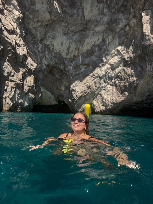 A person snorkeling in clear turquoise water near a large rocky cliff with a dark cave opening.
