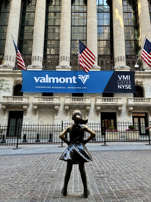 The image features a statue of a girl standing in front of a building, which has a sign that says "VALMONT V". The girl appears to be wearing a dress and holding her hands up in the air with her hair pulled back into a ponytail. There are also American flags flying above the building. In the background, there is another building with an advertisement for "chainlink" on it.