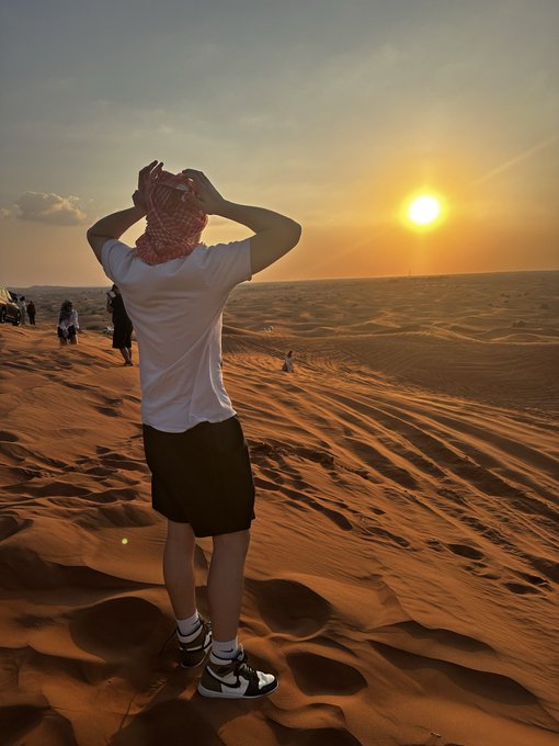 A person wearing a white t-shirt, black shorts, and a red keffiyeh stands on sand dunes in a desert, facing a sunset with the sun low on the horizon.