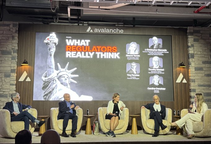 A panel of five people sit in chairs on a stage at an Avalanche event discussing "WHAT REGULATORS REALLY THINK," with panelists Tony Parsine, J. Christopher Giancarlo, and Caroline Crenshaw pictured on the screen.