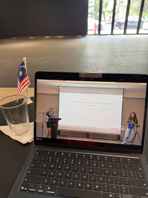 A laptop displays a presentation titled 'A calm place to save, spend and learn together,' with a small Malaysian flag placed in a glass beside it.