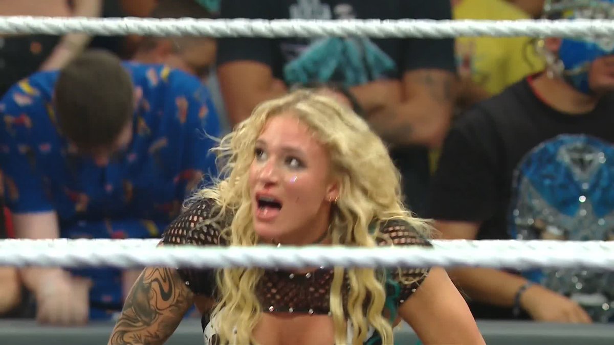 A blonde female wrestler with glitter on her face makes an excited, open-mouthed expression while leaning over the ropes of a wrestling ring.