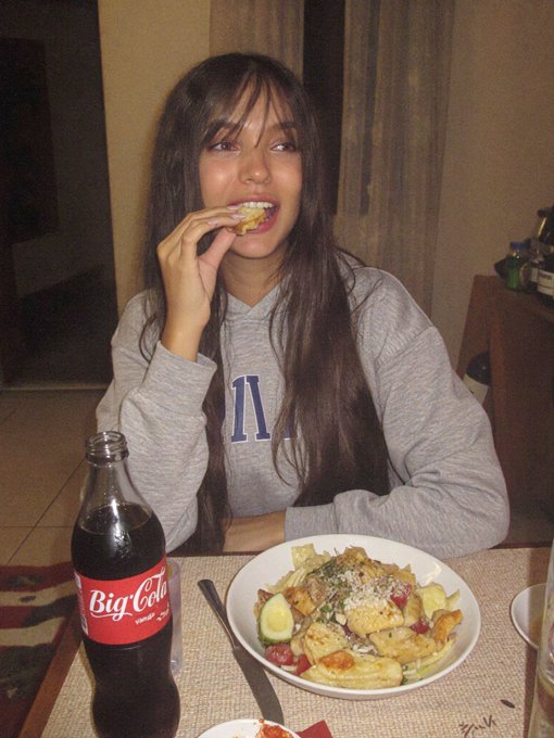 A woman in a gray sweatshirt sitting at a dinner table eating bread, with a bowl of pasta and a large Big Cola bottle in front of her.