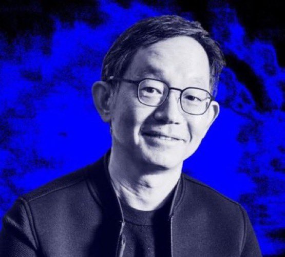 A black and white portrait photograph of a smiling man wearing glasses against a blue background.
