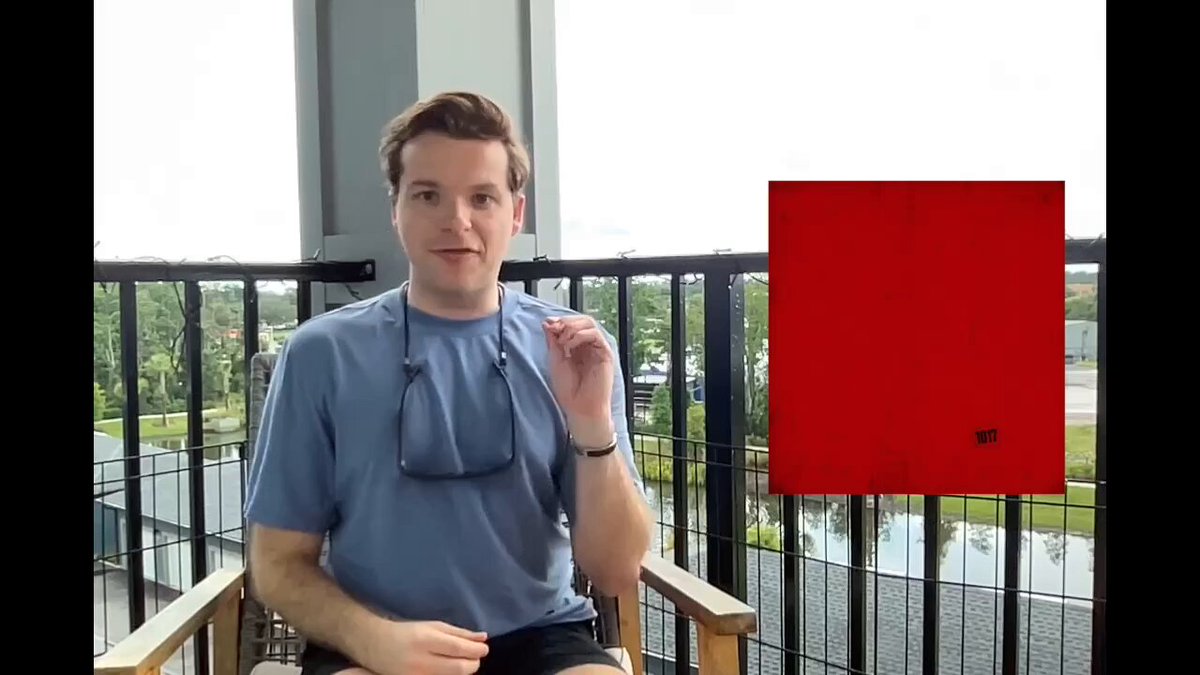 The image features a man sitting on a chair with his hand raised above his head. He is wearing a blue shirt, black shorts, and sunglasses. There are two red signs in the background of the image - one is larger than the other. The text "RED, WHITE, AND BORED" can be seen in the image as well. The man appears to be posing for a photo or participating in an online event related to the @BoredApeYC Confirms ApeFest 2026.