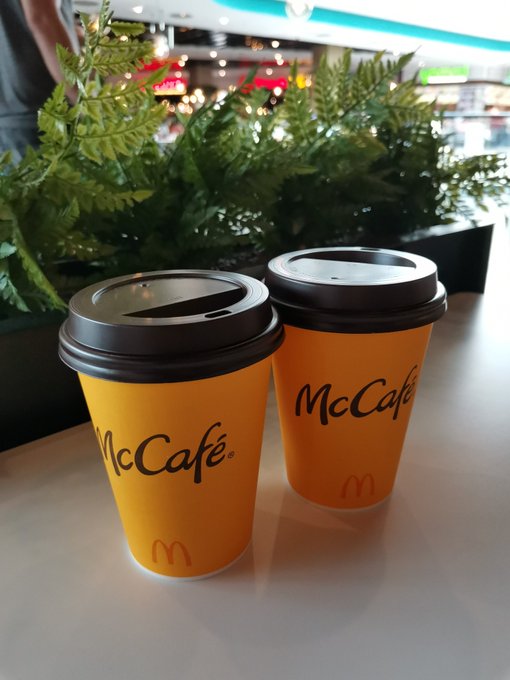 Two orange McCafé paper coffee cups with black lids sitting on a counter in what appears to be a mall or food court setting.