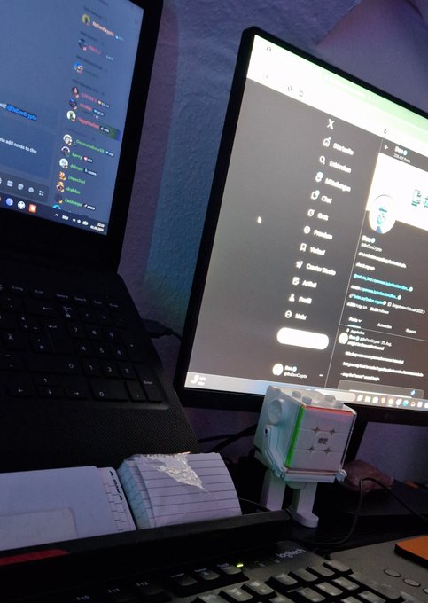 A desk setup with a laptop on the left showing a Discord chat, and a larger monitor on the right displaying the X (formerly Twitter) interface, with a small Cube Bot figure sitting between them on the desk.