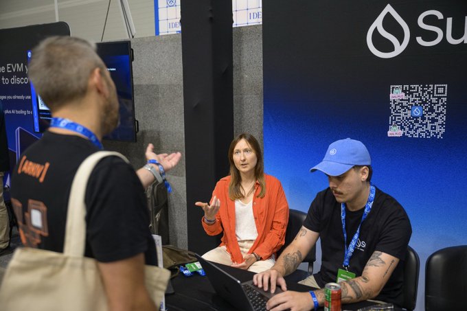 A man with a mustache and a woman in an orange jacket converse at the Sui Network booth during ETHGlobal Lisbon, as a Sui representative wearing a blue cap demonstrates something on a laptop displaying the venue's QR code. The branded blue backdrop and QR code promote Sui's developer tools (Move, Walrus, Seal, DeepBook, zkLogin) and the $6,000 in prizes referenced in the accompanying sponsorship post.