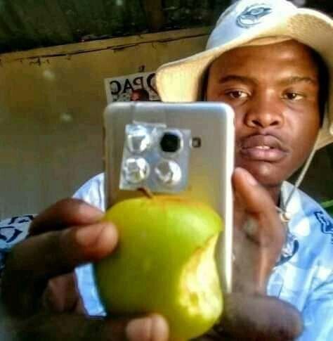 A man wearing a hat takes a mirror selfie with a smartphone, holding up a green apple to cover the camera area of the phone. The image humorously illustrates the post's joke about claiming to own an iPhone 17 while actually using an Android device, with the makeshift camera setup spoofing Apple's premium multi-lens design.