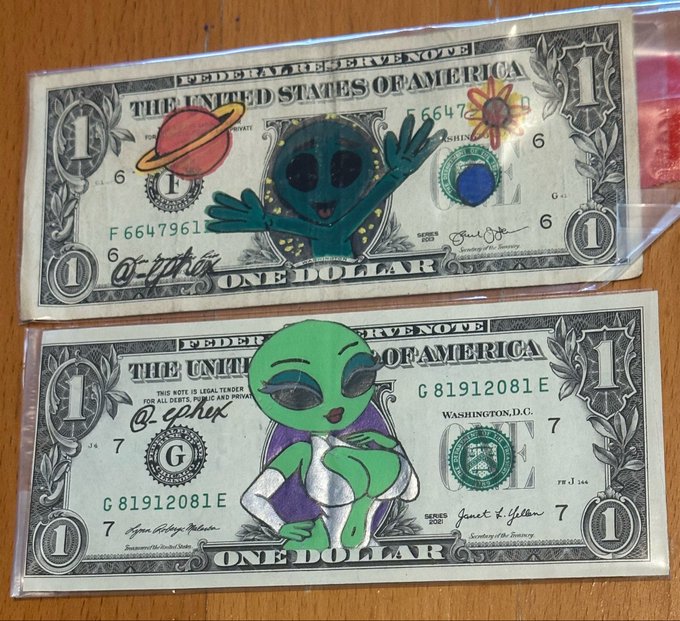 Two US one-dollar bills decorated with hand-drawn green aliens and planets, kept in protective sleeves.