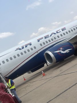 An Air Peace Boeing aircraft sits on the tarmac with a ground crew member standing nearby beside traffic cones, visually grounding the post's speculation about the Nigerian airline potentially adopting digital currency payments like Pi by showing the carrier at the center of the discussion. The visible "AIR PEACE" branding ties the airliner directly to the @flyairpeace mention in the accompanying text.