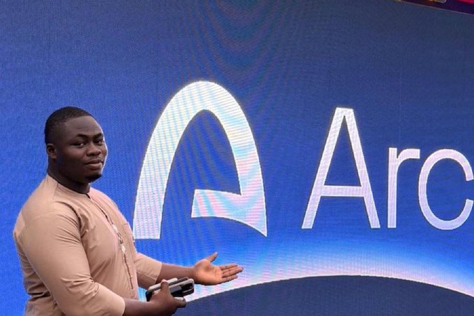 A man in a beige shirt gesturing while standing in front of a large blue display screen showing the Arc logo and partial 'Arc' text, appearing to give a presentation.