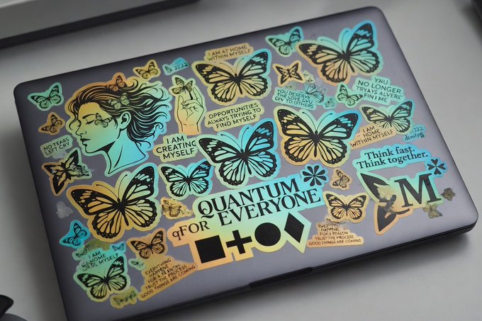 A laptop covered in holographic, iridescent stickers featuring butterflies, a woman's profile, geometric shapes, and motivational text including 'QUANTUM FOR EVERYONE' and 'Think fast, Think together.'