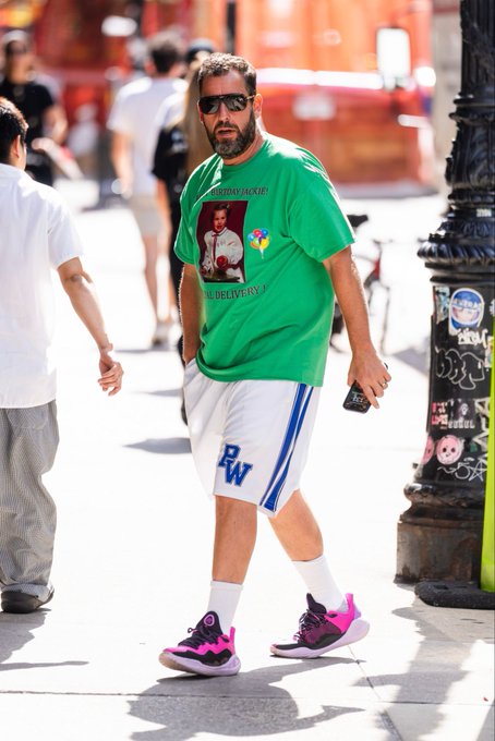 Adam Sandler walking on a city sidewalk wearing sunglasses, a green graphic t-shirt, white 'BW' shorts, and black and pink sneakers.