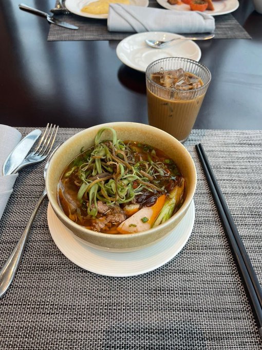A bowl of noodle soup with green onions and meat, accompanied by iced coffee, chopsticks, and a spoon, served on a dark wooden table with a placemat.