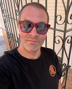 A selfie of a man wearing red-framed sunglasses and white earbuds, dressed in a black t-shirt with a small orange graphic, standing outdoors in front of a wrought-iron gate.