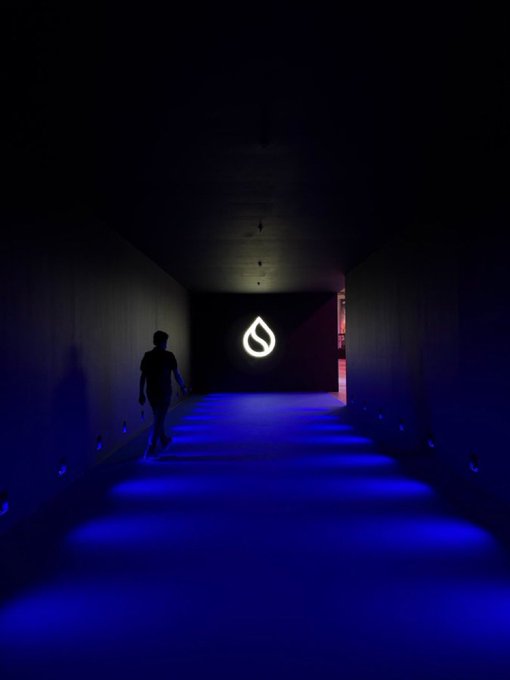 A person walks down a darkened corridor illuminated by blue LED lights on the steps, heading toward a glowing white flame or teardrop-shaped logo at the end of the hallway.