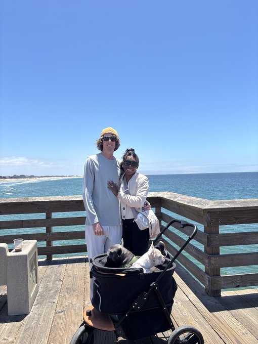 A couple stands on a wooden pier by the ocean with their two dogs in a pet stroller; the man wears a yellow beanie and light blue shirt while the woman wears sunglasses and a beige cardigan.