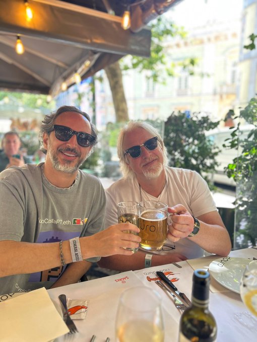 The image shows two men sitting at a table, holding glasses of beer together as they pose for a picture. The table also has wine glasses, a bottle, and utensils like forks and knives. In the background, there are potted plants adding greenery to the scene. The post in the image is about celebrating an event or occasion with friends, possibly a date or a party.