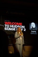 A woman presenting on stage in front of a screen displaying "WELCOME TO HUDSON STAGE 2" with her name "Paige," captured in a vertical, slightly blurry photo.
