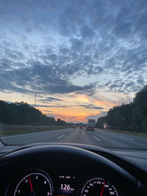 View from inside a car driving on a highway at sunset, with the dashboard showing a speed of 126 km/h and dramatic cloudy sky overhead.