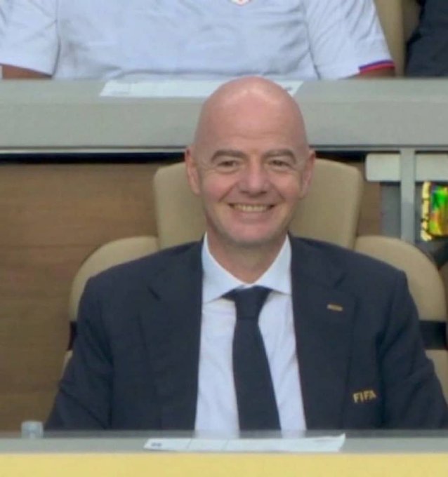 A bald man in a dark suit and black tie smiles while seated in what appears to be a VIP box, with a FIFA-branded item visible nearby.