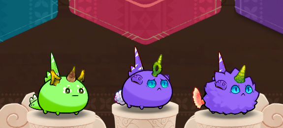 Three colorful cartoonish axolotl-like creatures (green and two purple ones) wearing party hats, sitting on pedestals in what appears to be a game screenshot.