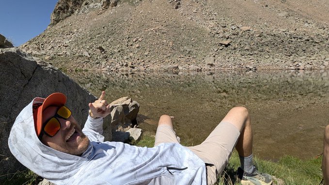 A man wearing a hooded sun shirt, sunglasses, and a cap lies on the ground beside a mountain lake, pointing up at the rocky cliff with a smile. The relaxed outdoor setting complements the post's theme of self-custody and home mining, evoking a sense of freedom and independence that aligns with the crypto self-sovereignty message from CryptoCloaks.com.