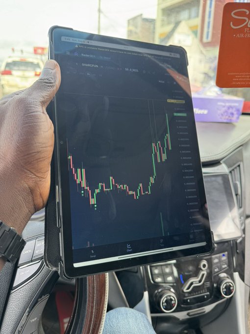 A person holds up a tablet inside a car displaying a candlestick chart for "$SHARCFUN" with a notable upward price spike, visually supporting the post's call to send the meme token "higher."