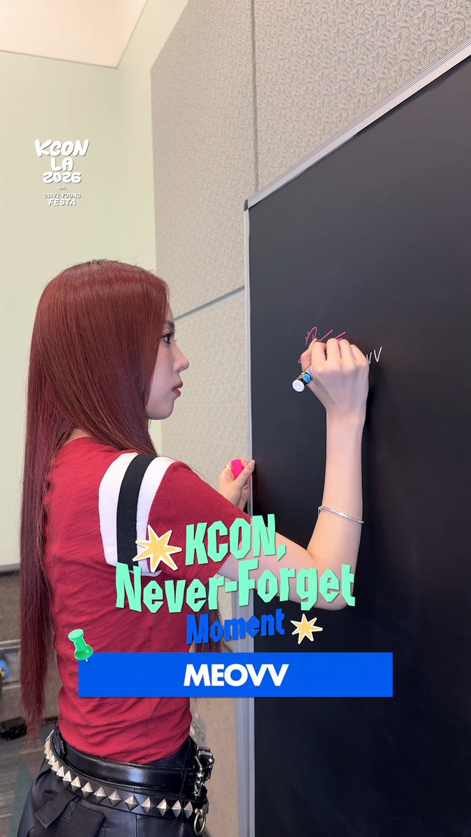 A red-haired member of MEOVV signs a black board in pink marker at KCON LA 2026, with the "KCON Never-Forget Moment" graphic highlighting the group's featured appearance at the event.