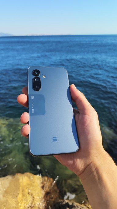 A hand holds a light blue Meta Vault smartphone with 'META VAULT' branding on the back, displayed against a clear blue sea and sky.