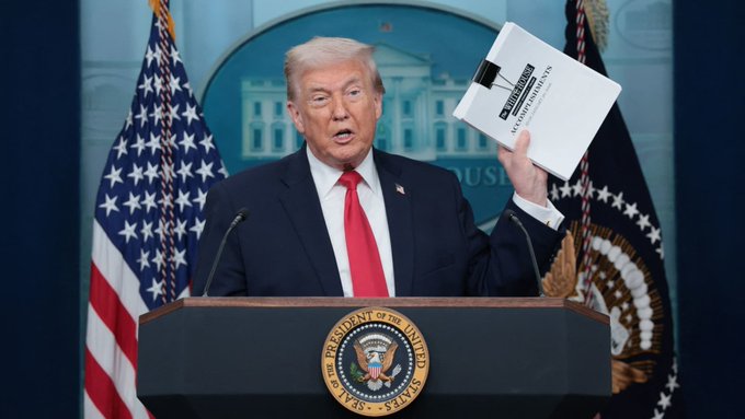Donald Trump holds up a binder labeled "The White House Accomplishments" while standing at a podium. The image relates to the post by highlighting a past administration's focus on achievements, while the current post discusses delays in a new initiative.