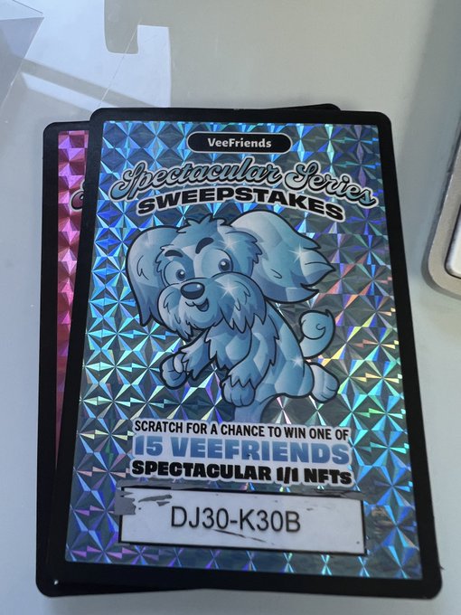 A holographic VeeFriends Spectacular Series Sweepstakes card featuring a cartoon dog, with a code 'DJ30-K30B' at the bottom, advertising a chance to win one of 15 VeeFriends Spectacular 1/1 NFTs.