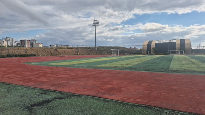 An empty sports complex features a red running track surrounding a green turf field, with a large modern building labeled 'NEXT ONE' visible on the right under a cloudy sky.