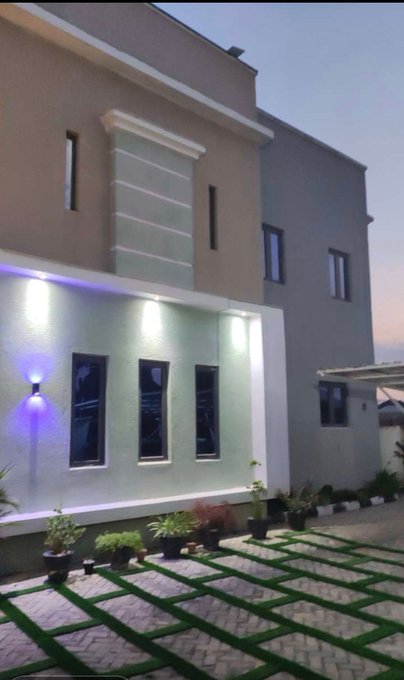 A modern two-story villa at dusk is illuminated by accent lighting, featuring a paved courtyard with grass-filled grid patterns and potted plants lining the walkway, with no visible text or numbers. The image does not relate to the post, which discusses financial struggles ("profit," "Notcoin & memefi Influencer," "Tinubu Regime") that are unrelated to a real estate property photo.