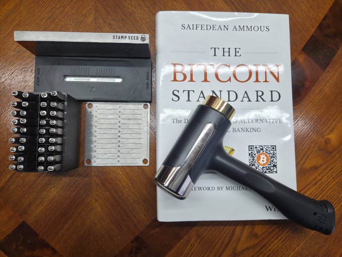 A wooden table displaying 'The Bitcoin Standard' book by Saifedean Ammous alongside a power drill and a stamp seed kit containing letter punches and plates.