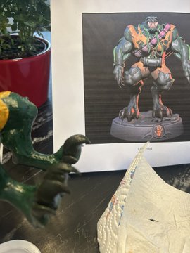 A green ceramic figurine of a reclining figure sits next to a framed picture of a monstrous, muscular character with glowing red eyes, displayed on a wooden surface near a potted plant.