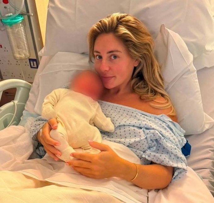 A smiling woman in a hospital gown holding a newborn baby (whose face is blurred) while resting in a hospital bed.