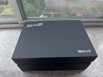 A sleek black OKX VIP gift box labeled "NEW MONEY HAPPENING HERE" sits on a windowsill, showcasing the premium merchandise the user received from the cryptocurrency exchange as part of their World Cup-themed peripheral collection. The image illustrates the tweet's appreciation for OKX's thoughtful giveaway while teasing the user about collecting similar merchandise from other exchanges.