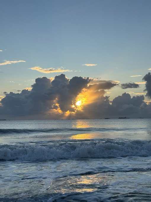 Sunbeams break through dramatic cumulus clouds at sunset, casting a golden reflection across ocean waves rolling onto a beach.