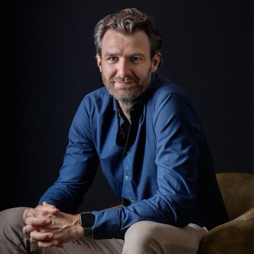 Professional portrait of a bearded man in a blue button-down shirt and khaki pants, seated with hands clasped against a dark background.