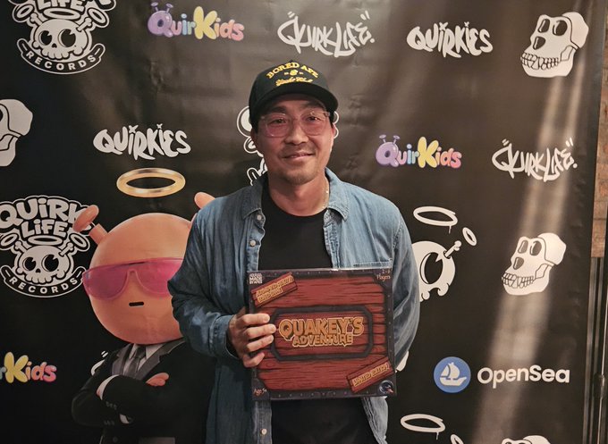 A man wearing glasses and a cap poses with a 'Quirkeys Adventure' collectible box at a Quirk Life / Quirkeys event, with OpenSea branding visible.