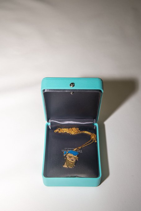 A turquoise Tiffany & Co. jewelry box is open to reveal a gold pendant on a chain, featuring what appears to be a CryptoPunks design, displayed against a dark interior with partial "TIFFANY & CO." branding visible. The image showcases the never-worn CryptoPunks x Tiffany & Co. pendant (1/250) that the seller is parting with to fund photography expenses.