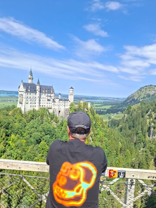 A person wearing a black t-shirt featuring an orange boxing glove design and a black cap, looking out over a valley towards a large white castle (Neuschwanstein) in Bavaria.
