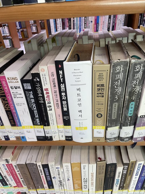 A library bookshelf displays Korean-language books alongside English titles like "Bitcoin: A Peer-to-Peer Electronic Cash System" (by Satoshi Nakamoto's white paper) and "The BlockSize War," with the image humorously implying the poster is still "reading up" rather than investing, suggesting retail investors remain in research/observation mode before committing seed money to trigger the crypto bull market.