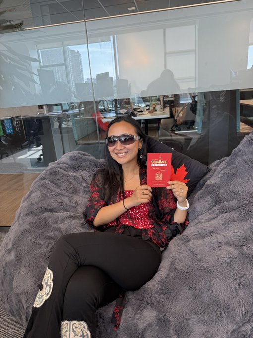 A smiling woman wearing sunglasses sits on a gray fluffy beanbag in a modern office space, holding a red card that appears to be a Permanent Resident card with a Canadian maple leaf design.
