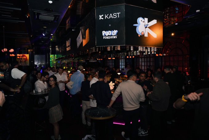 A dimly lit nightclub scene with a crowd of people socializing, featuring illuminated signs displaying 'KAST x Pudgy Penguins' branding hanging from the ceiling.