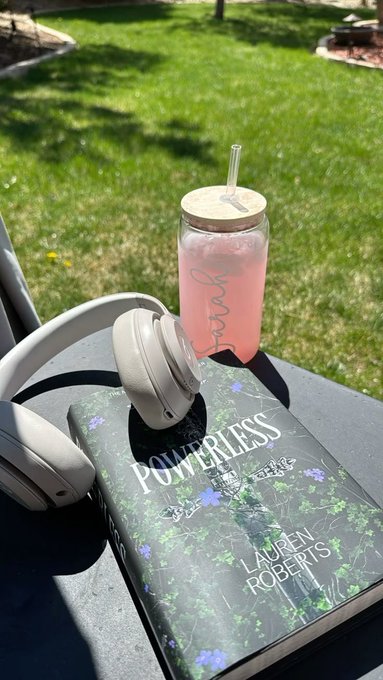 A pink smoothie in a 'Love' branded glass bottle with a straw sits on a book titled 'POWERLESS' by Lauren Roberts, accompanied by white headphones on a table outdoors.